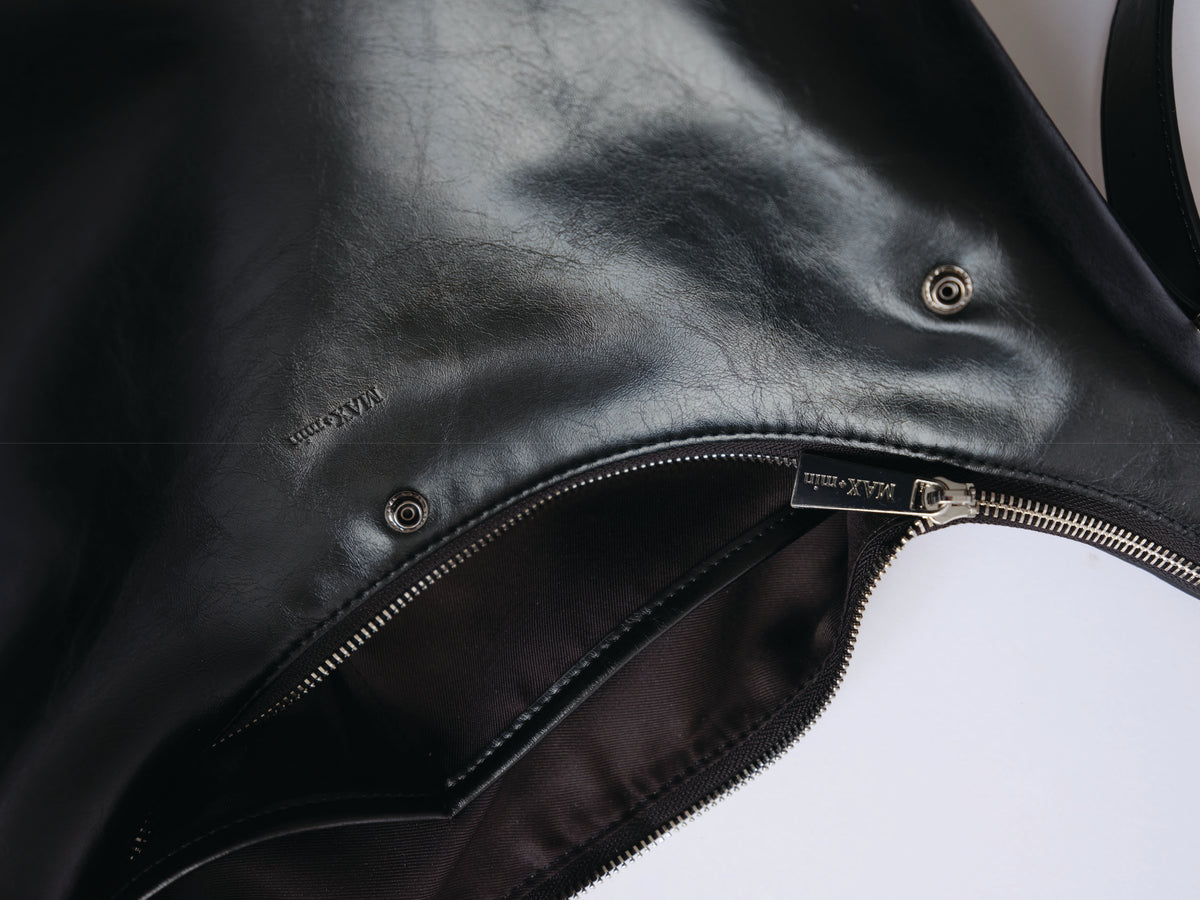 Top detail of the MAX+min Maximum Bag showing black full-grain leather, zipper closure, and MAX+min hardware.